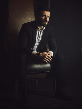 A handsome man with a beard and in a classic suit sits in a leather chair and looks at the camera. 