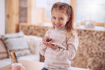 Modern device. Joyful cute girl smiling while using a modern tablet