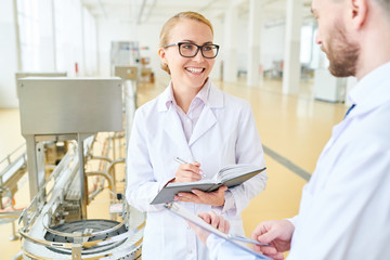 Obraz premium Portrait shot of attractive technologist with charming smile talking to male colleague and taking notes while having working meeting at spacious production department of milk factory.