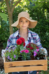 Beautiful female gardener wearing straw hat holding wooden crate full of flowers ready to be planted in her garden. Gardening concept.