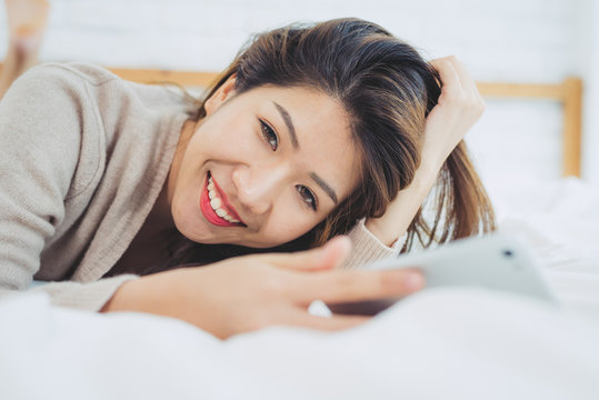 Happy Asian women are using smart phone on the bed in morning. Asian woman in bed checking social apps with smartphone. Smiling woman surfing net with cellphone at home. Mobile addict concept.