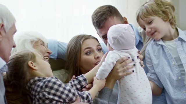 PAN of happy young woman holding cute baby girl and cooing over her while sitting on sofa with excited family of husband, grandparents and children