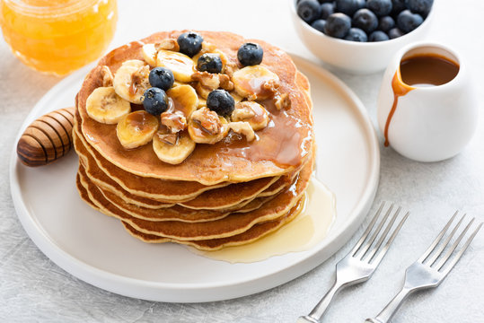 Stack of pancakes with banana, blueberries, walnuts, honey and caramel sauce on white plate. Closeup view. Breakfast pancakes with maple syrup