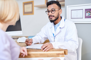 Portrait of handsome Middle-Eastern doctor consulting young woman sitting at desk in modern office, copy space