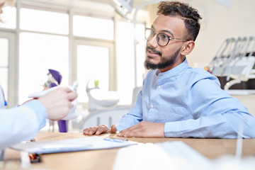 Portrait of modern Middle-Eastern man talking to dentist sitting at desk in modern dental clinic, copy space