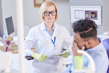 Portrait of smiling female dentist listening to patient sitting in dental chair and complaining about toothache in modern dental clinic, copy space