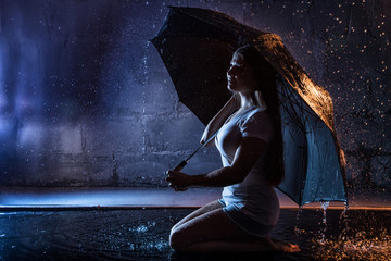 Girl in the white shirt with black umbrella, water drops around and dark walls background illuminated by light during a photoshoot with water