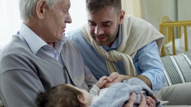 Medium shot of young father touching hand of baby sleeping in arms of grandfather