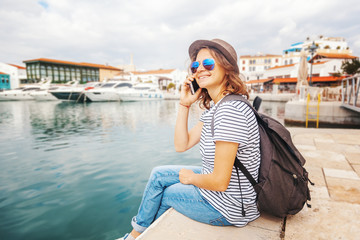 Fototapeta na wymiar young beautiful woman traveler in a hat talking on a mobile phone on a beautiful port background on a sunny day by the sea. Vacations, adventures and sea voyages