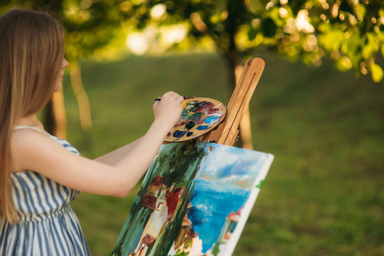 Beautiful girl standing in the park and draws a picture using a palette with paints and a spatula.