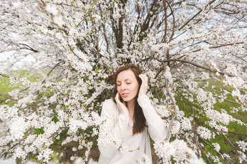 Young relaxed tender beautiful woman in light casual clothes keeping hands near face standing in city garden or park on blooming tree background. Spring nature, flowers. Lifestyle, leisure concept.