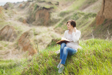 Young smiling relaxed woman in light casual clothes sitting on grass studying reading book on green field background. Student learning, education. Beautiful landscape. Lifestyle, leisure concept.