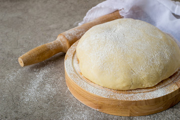 Homemade dough on a round wooden board, next to a rolling pin and a napkin made of cotton white fabric. 