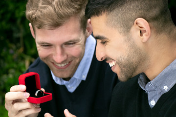 Engagement proposal betwen two gay men as one man proposes with an engagement ring in red box
