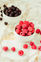 Raw raspberries and wild black, blackberries in white cups. Light marble background. Summer fruit, berries. Detox diets and healthy food concept.