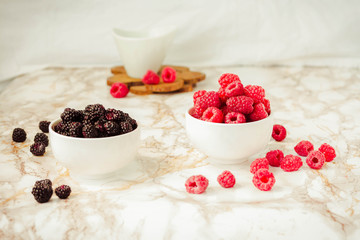 Raw raspberries and wild black, blackberries in white cups. Light marble background. Summer fruit, berries. Detox diets and healthy food concept.