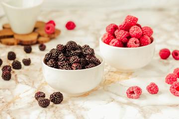 Raw raspberries and wild black, blackberries in white cups. Light marble background. Summer fruit, berries. Detox diets and healthy food concept.