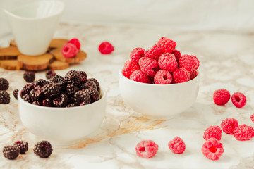 Raw raspberries and wild black, blackberries in white cups. Light marble background. Summer fruit, berries. Detox diets and healthy food concept.