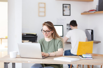 Successful smiling businesswoman dressed in casual clothes, sits at work place, studies documents, surfing high-speed internet on laptop pc for reading news. Coworking and startup concept