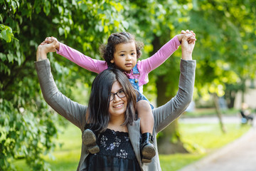 Mixed race mother carrying excited hispanic toddler daughter three years old in blue denim sarafan and pink shirt holding on her shoulders while walking together in summer park.