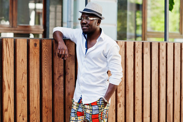 Stylish african american man in white shirt and colored pants with hat and glasses posed outdoor. Black fashionable model boy.