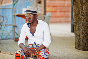 Stylish african american man in white shirt and colored pants with hat and glasses posed outdoor. Black fashionable model boy.