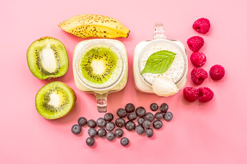 two banana and kiwi milkshakes in mason jars with creme on top decorated with kiwis, bananas, raspberries and blueberries. flat lay top view smoothie on pink fashion background