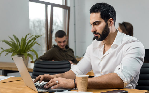 Attractive Bearded Man In Pristine White Shirt Working On Silver Laptop In Modern Office