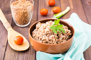 Cooked buckwheat in a bowl with a sheet of parsley, a glass of raw buckwheat and cherry tomatoes on a wooden table