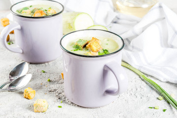 Two portions of homemade zucchini creamy soup with bread crumbs in mugs on a light concrete background.