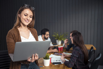 Pretty businesswoman using laptop as standing in the office, three smart managers discussing possible solutions at the desk in the background, portrait shot