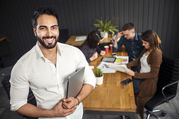 Fototapeta premium Pleased asian man with black beard holding silver laptop, standing before colleagues working on the task, preparing next stage of the project at office desk with graphs, coffee cups and gadgets