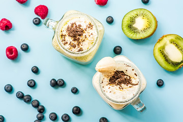 two banana and kiwi milkshakes in mason jars with creme and chocolate chips on top decorated with kiwis, raspberries and blueberries scatter. flat lay top view smoothies on blue fashion background