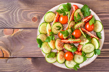 Vegetable salad of fresh cucumbers, tomatoes, parsley and sesame seeds on a plate on a wooden table. Top view