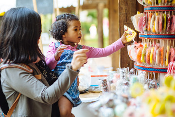 Mexican mother with dark skinned hispanic toddler girl choosing lollipop in candy showcase in open...