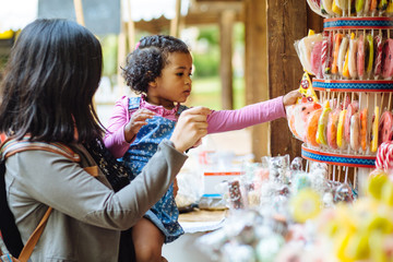 Mexican mother with dark skinned hispanic toddler girl choosing lollipop in candy showcase in open...