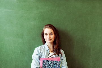Young confident smiling female high school student standing in front of chalkboard in classroom, holding textbooks, looking at camera. Waist up portrait.