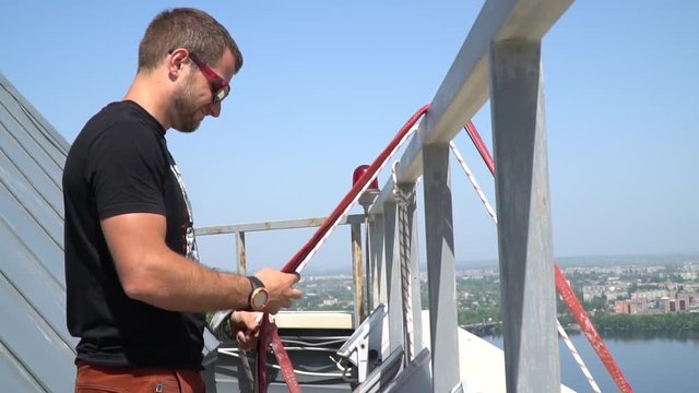 A man in sunglasses stands on the roof of the building and prepares slings for the slack line