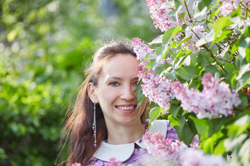 Naklejka premium A smiling young woman with black hair peeks out from behind the flowers of purple lilac 