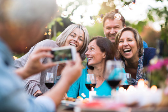 Some friends in their 40s gathered around a table in the garden to share a meal. A man takes a picture of a group of friends
