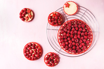 Variety of red raspberry shortbread tarts and tartlets with lemon custard and glazed fresh raspberries served on cooling rack over pink pastel background. Top view, space.