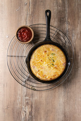 Spanish potato omelette tortilla with bacon served in cast-iron pan with tomato sauce on metal cooling rack over wooden background. Top view, space.