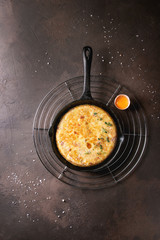 Spanish potato omelette tortilla with bacon served in cast-iron pan on cooling rack with egg yolk and salt over over dark brown texture background. Top view, space.