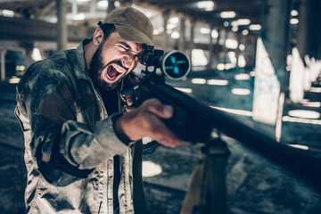 Fighter is looking through lens on rifle and taking aim. He is holding it with both hands. Emotional guy is screaming and yelling. He is in a big hangar alone.