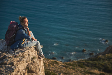 Fototapeta na wymiar Glad female tourist communicating by cellphone while sitting on the top of mounting by the sea. Copy space in right side