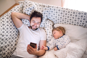 Father with smartphone and a sleeping toddler boy in bed at home.
