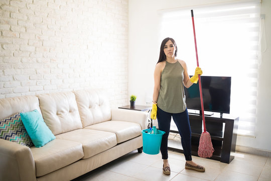 Stressed woman standing holding broom and bucket in living room