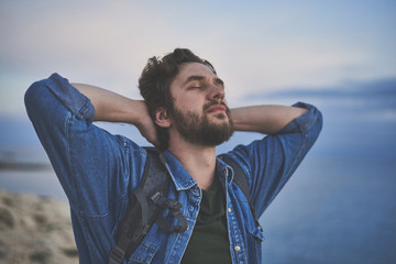 Portrait of relaxed young man enjoying sea breeze with closed eyes. He is standing with backpack