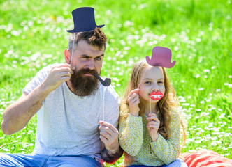 Dad and daughter sits on grass at grassplot, green background. Aristocrates concept. Child and father posing with top hat, lips and pipe photo booth attributes. Family spend leisure outdoors.