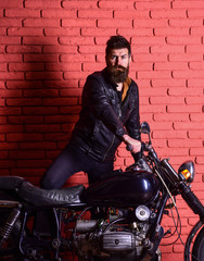 Hipster, brutal biker on serious face in leather jacket gets on motorcycle. Man with beard, biker in leather jacket near motor bike in garage, brick wall background. Masculine passion concept.